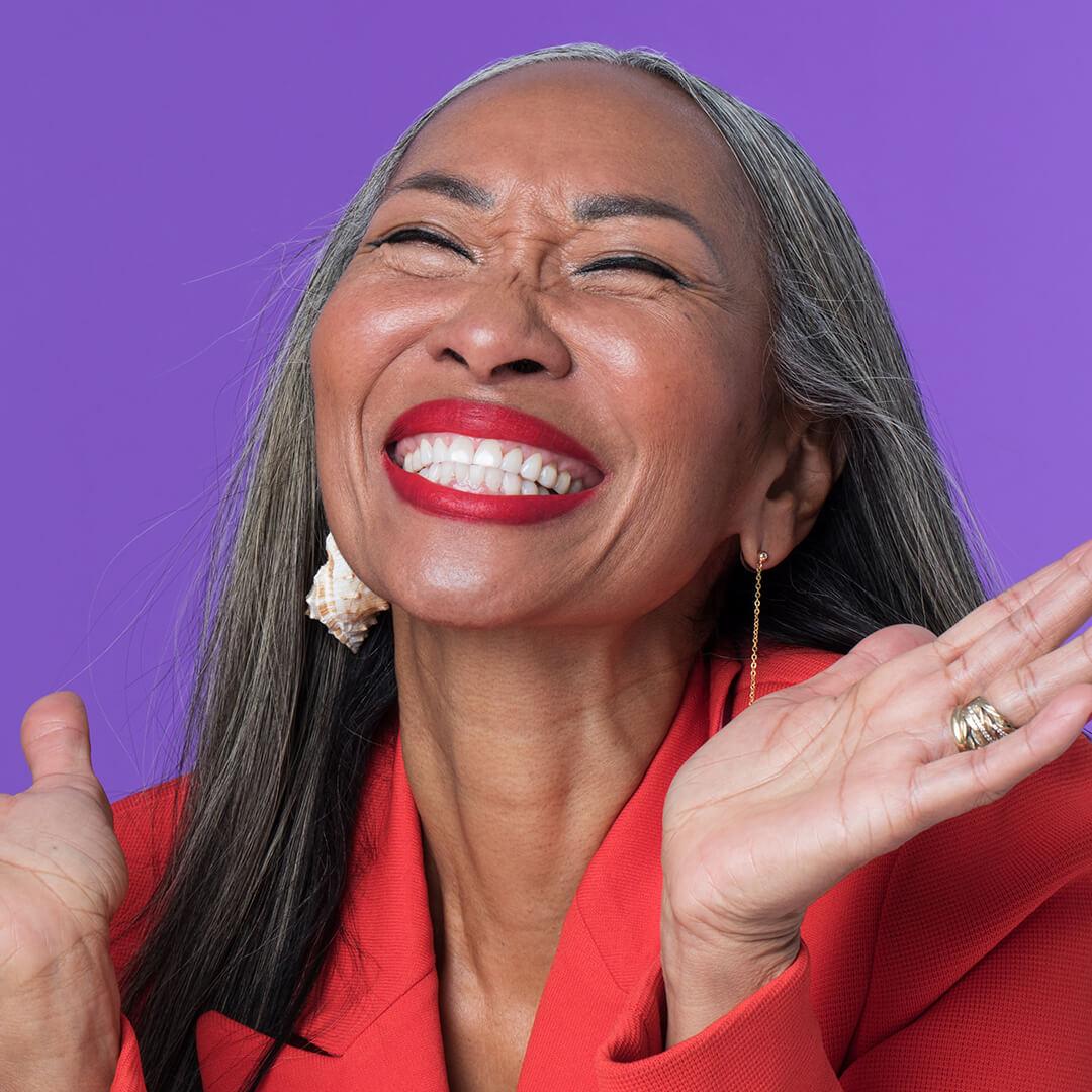 A photo of a mature woman laughing wearing red lipstick and sporting a red coat and conch earrings on a purple background Desktop