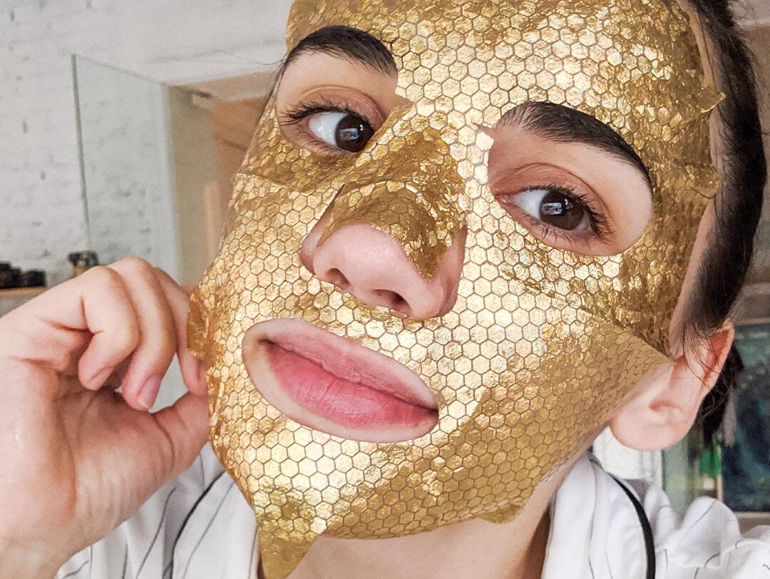 Photo of a young woman in her apartment putting on a gold sheet mask