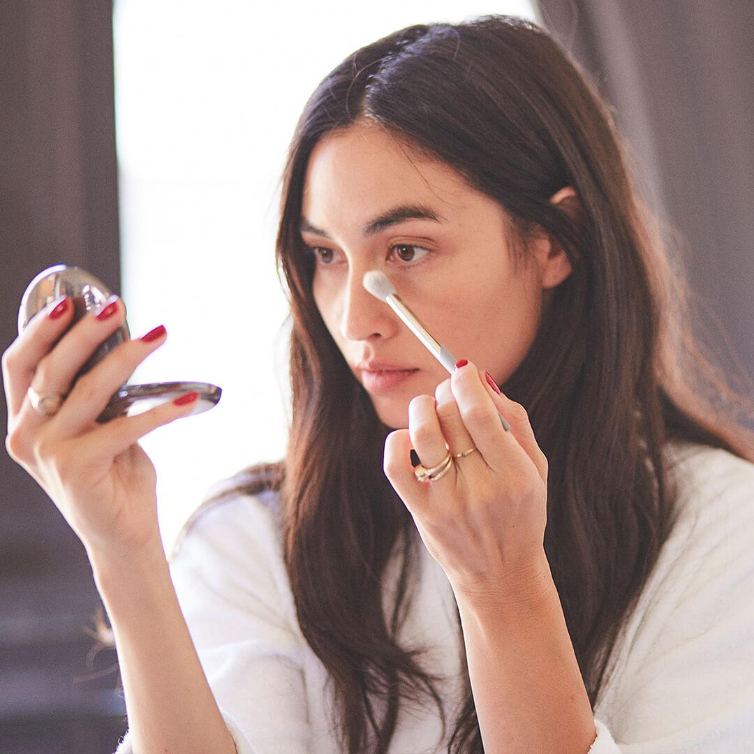 Beautiful young model contouring her nose using a COMPLEX CULTURE makeup brush and holding a compact mirror Mobile